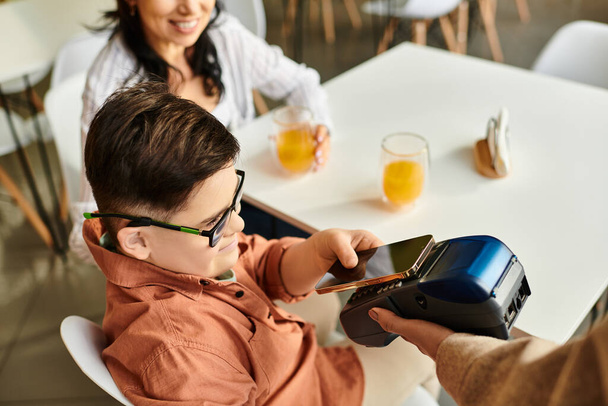 adorable inclusive chłopiec z zespołem Downa płacąc smartfonem w kawiarni w pobliżu jego wesoła matka - Zdjęcie, obraz