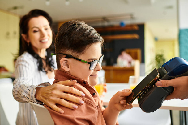 rozkošný inkluzivní chlapec s Downovým syndromem platící smartphonem v kavárně poblíž jeho veselá matka - Fotografie, Obrázek