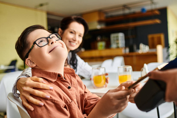 suloinen inclusive poika Downin oireyhtymä maksaa älypuhelimella kahvilassa lähellä hänen iloinen äitinsä - Valokuva, kuva