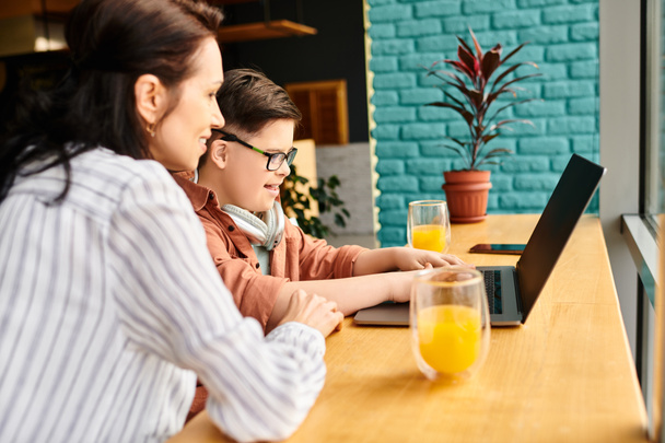веселый инклюзивный мальчик с синдромом Дауна с наушниками, играющий на ноутбуке рядом с матерью в кафе - Фото, изображение