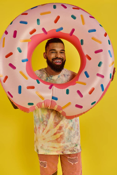 Ein Mann versteckt spielerisch sein Gesicht hinter einem massiven Donut und zeigt sich von seiner skurrilen und humorvollen Seite, während er ein Leckerli genießt. - Foto, Bild