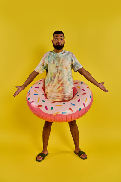 Een stijlvolle man in een tie-dye shirt houdt een kleurrijke donut float, staande te midden van een zomers tafereel met een grillige touch. - Foto, afbeelding