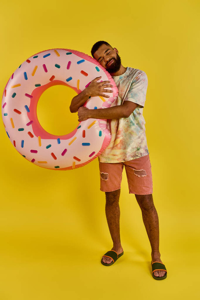 Een man houdt vrolijk een gigantische donut voor een levendige gele achtergrond, en toont zijn liefde voor de zoete traktatie. - Foto, afbeelding