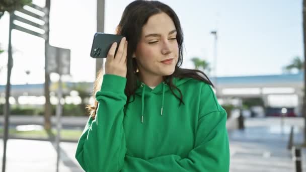 Jovem atraente com capuz verde sorri enquanto ouve uma mensagem de voz em seu smartphone ao ar livre na cidade. - Filmagem, Vídeo