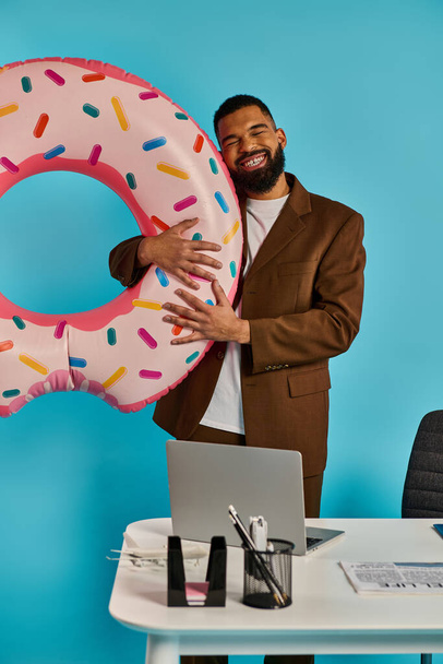 Мужчина держит огромный пончик перед ноутбуком, похоже, взаимодействуя с экраном. Сопоставление сладкого удовольствия и технологий создает причудливую и сюрреалистичную сцену.. - Фото, изображение