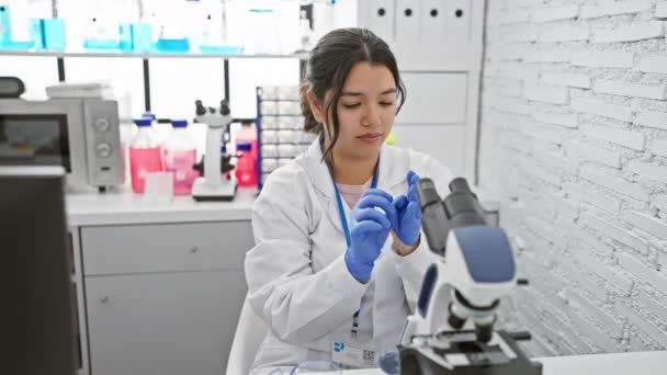 Una joven científica hispana examinando muestras con un microscopio en un moderno laboratorio. - Metraje, vídeo
