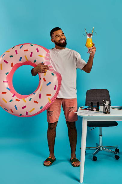 Ένας άντρας με χαρά κρατάει ένα ποτό και ένα τεράστιο ντόνατ στα χέρια του, απολαμβάνοντας ξεκάθαρα τις απολαύσεις του.. - Φωτογραφία, εικόνα