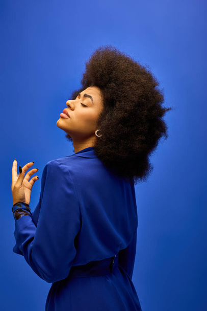 Une femme afro-américaine à la mode avec une coiffure bouclée pose dans une robe bleue sur un fond coloré. - Photo, image