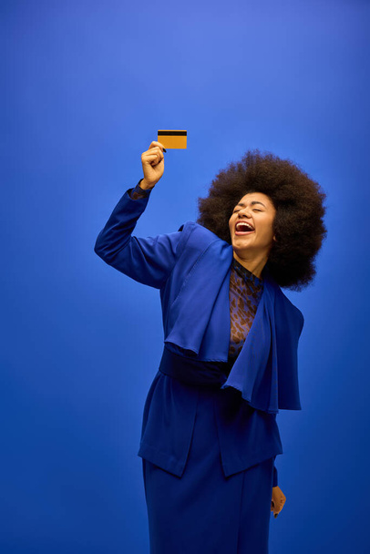 鮮やかな背景に対して青いスーツでクレジットカードを保持するスタイリッシュなアフリカ系アメリカ人女性. - 写真・画像