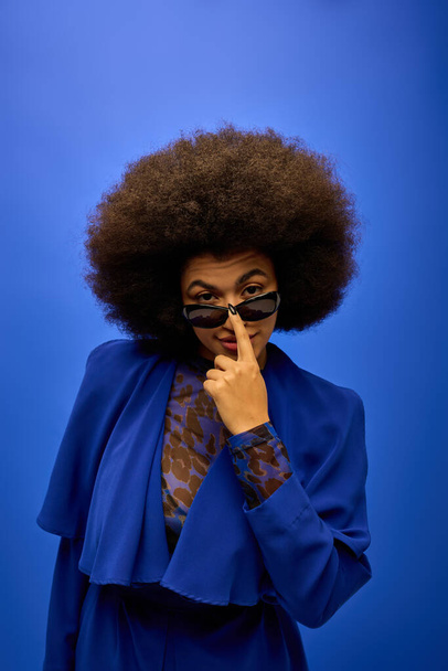 Stijlvolle Afro-Amerikaanse vrouw poseren in trendy blauwe jas en bril op levendige achtergrond. - Foto, afbeelding