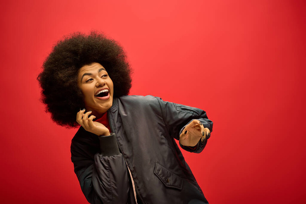 Mujer afroamericana con un impresionante peinado afro de pie con confianza sobre un fondo rojo audaz. - Foto, imagen