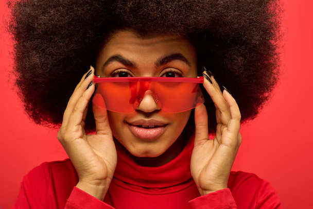 Stylowa Afroamerykanka z kręconą fryzurą trzyma parę czerwonych okularów. - Zdjęcie, obraz