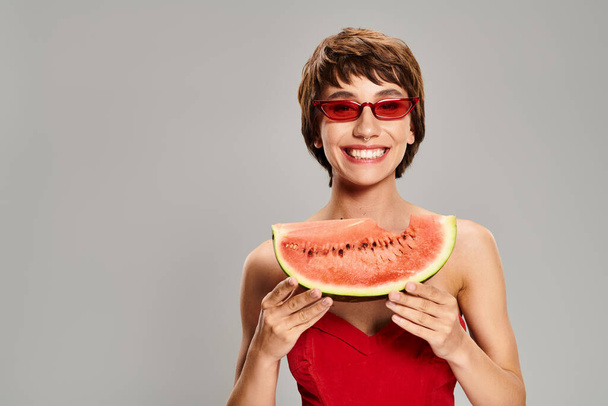 Een stijlvolle jonge vrouw in een rode jurk houdt heerlijk een plakje watermeloen vast. - Foto, afbeelding
