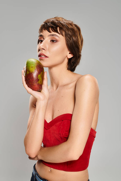 Eine junge Frau in rotem Oberteil hält eine reife Mango in einer lebendigen Umgebung. - Foto, Bild