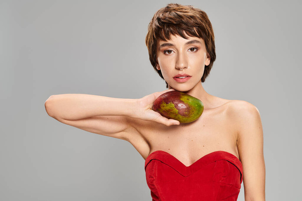 Eine hinreißende junge Frau steht elegant in einem roten Kleid und hält eine lebendige Mango in der Hand. - Foto, Bild