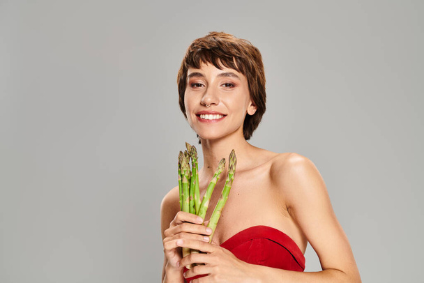 A captivating young woman in a radiant red dress holds a bunch of fresh asparagus. - Photo, Image