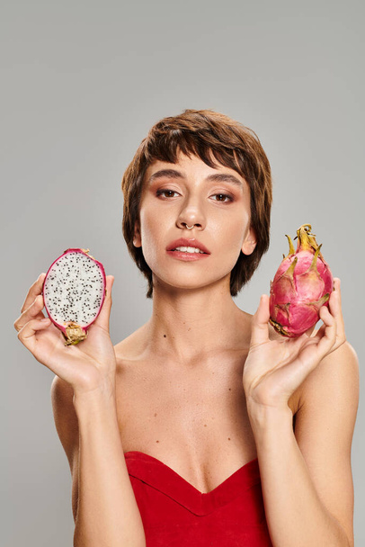 A stunning woman in a red dress gracefully holds a ripe dragon fruit. - Photo, Image