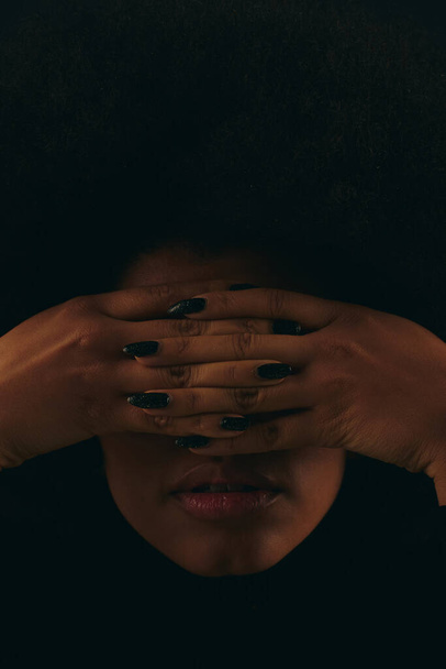 Μια κομψή Αφροαμερικανή με μοντέρνα ενδυμασία καλύπτει το πρόσωπό της με τα χέρια της. - Φωτογραφία, εικόνα