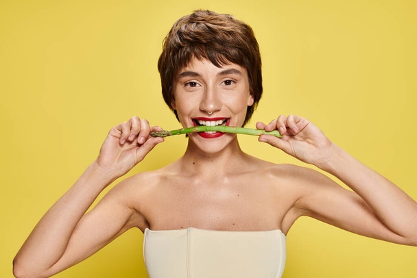 Młoda kobieta trzyma szparagi w pobliżu ust, na tętniącym życiem tle. - Zdjęcie, obraz