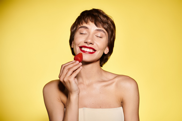 Жінка делікатно тримає полуницю на обличчі на яскравому тлі. - Фото, зображення