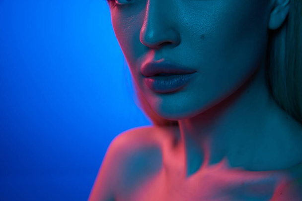 Cropped close-up image of beautiful female plump lips against blue studio background in neon light. Lip augmentation. Concept of natural beauty, cosmetology, cosmetics, skin care and health - Photo, Image