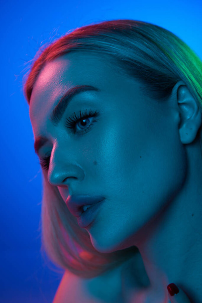 Portrait of attractive young woman with perfect, spotless skin posing against blue studio background in neon light. Concept of natural beauty, cosmetology, cosmetics, skin care and health - Φωτογραφία, εικόνα