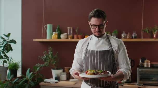 Retrato medio del chef masculino caucásico con uniforme de chefs y anteojos posando para la cámara con delicioso filete de carne y verduras a la parrilla en el plato - Imágenes, Vídeo