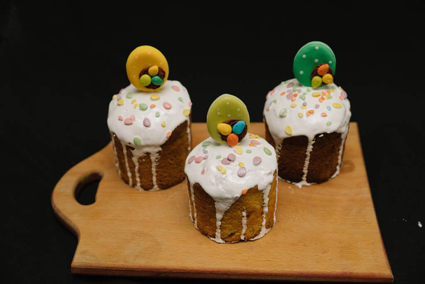 Velikonoční dorty s bílou polevou a barevné dekorace na dřevěné desce na černém pozadí. - Fotografie, Obrázek