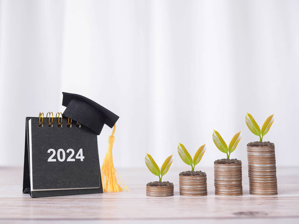 2024 ημερολόγιο γραφείο με καπέλο αποφοίτησης και τα φυτά μεγαλώνουν σε στοίβα των κερμάτων. Η έννοια της εξοικονόμησης χρημάτων για την εκπαίδευση, φοιτητικό δάνειο, υποτροφία, δίδακτρα στο μέλλον - Φωτογραφία, εικόνα