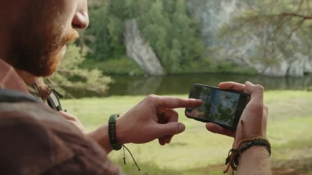 Közelkép ív lövés a férfi turista vizsgálja térképet okostelefon képernyőn, és rámutatva a hegyre, miközben túrázik a barátnőjével a természetben - Felvétel, videó