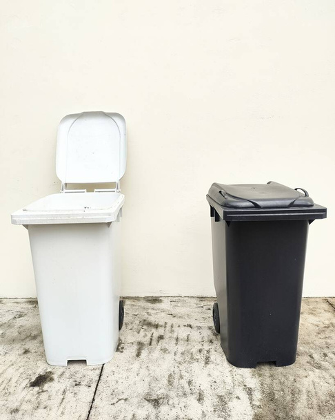 Een witte en een zwarte vuilnisbak naast elkaar voor een witte muur. Voorbeeld van beheer en recycling van stedelijk afval. - Foto, afbeelding