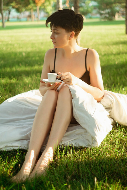 Ein in eine Decke gehülltes Mädchen sitzt mit einer Tasse Kaffee auf grünem Gras und genießt ein erfrischendes Morgengebräu. Coffeeshops im Park bieten einen herrlichen Start in den Tag mit belebendem Morgenkaffee. - Foto, Bild
