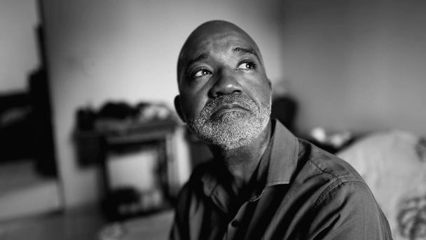 Продуманий депресивний афроамериканський старший чоловік з сумними емоціями бореться в самоті в мотивованій спальні в драматичному чорно-білому, монохромному портреті - Фото, зображення