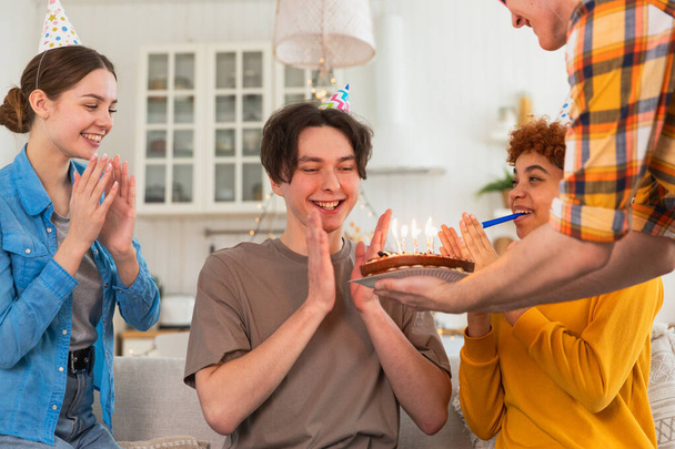 Bir dilek tut. Parti şapkası takmış bir adam doğum günü pastasında yanan mumları üflüyor. Doğum günün kutlu olsun. Arkadaş grubu adama mutlu yıllar diliyor. İnsanlar doğum gününü evde partiyle kutluyor. - Fotoğraf, Görsel