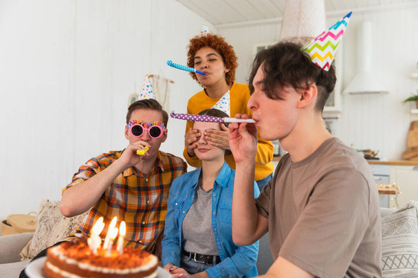 Fais un vœu. Femme portant une casquette de fête soufflant des bougies brûlantes sur le gâteau d'anniversaire. Joyeux anniversaire. Groupe d'amis souhaite un joyeux anniversaire à la fille. Les gens qui célèbrent leur anniversaire avec fête à la maison - Photo, image