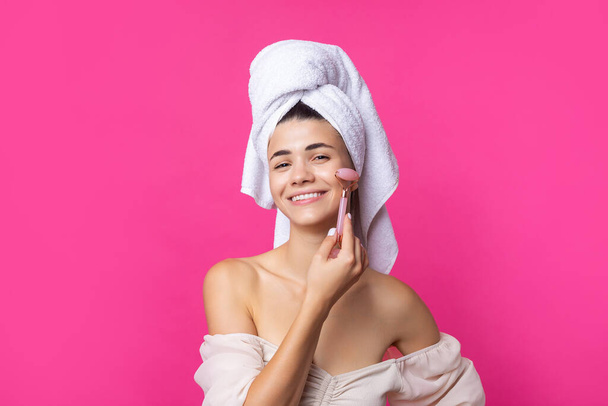 A beautiful cheerful attractive girl with a towel on her head holds a cosmetic roller near her face against a pink background. - Photo, Image