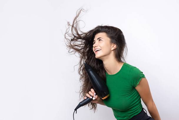A cute young girl dressed in a green top dries her beautiful long silky hair with a hair dryer against a white background. - Fotografie, Obrázek
