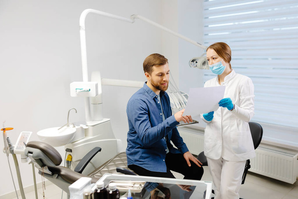 Νεαρός ελκυστικός άντρας που επισκέπτεται οδοντίατρο, κάθεται σε οδοντιατρική καρέκλα στη σύγχρονη κλινική φωτός. Νεαρή οδοντίατρος κρατώντας ακτινογραφία εικόνας - Φωτογραφία, εικόνα