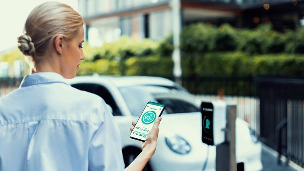 ビジネスウーマンは,エコグリーンシティパークの充電ステーションから充電しながら,スマートフォン画面でEVバッテリーステータスモバイルアプリケーションをチェックします. 電気自動車の未来的なエネルギーの持続可能性.Peruse - 写真・画像
