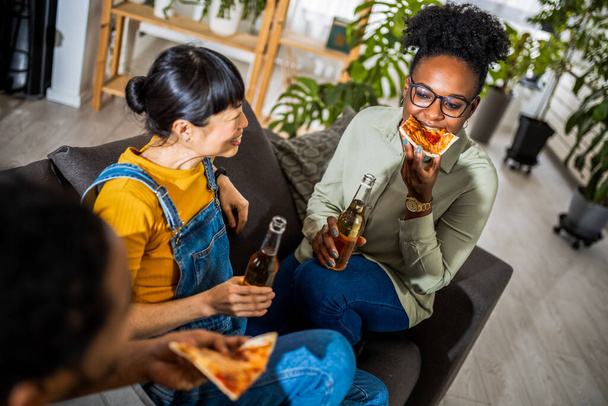 Мультиэтническая группа друзей пьет пиво и ест пиццу дома. - Фото, изображение