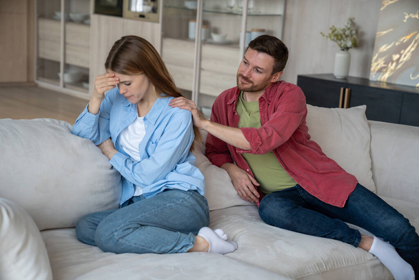 Difficulties family relationships reflected in posture offended woman sitting on home sofa. Wife pointedly turned away from husband. Man is trying to make peace gently puts hands on beloveds shoulders - Photo, Image