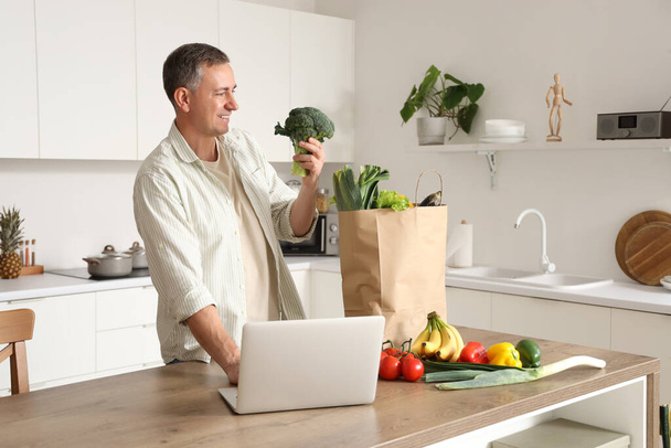 Homme mûr avec brocoli frais en utilisant un ordinateur portable dans la cuisine - Photo, image