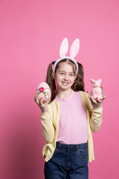 Joyful toddler presenting a pink egg and a stuffed rabbit in studio, wearing pigtails and cute bunny ears on camera. Smiling young kid showing her easter decorations and colorful toys. - Photo, Image
