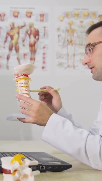 Teini-ikäinen ortopedien tapaamisessa. Ortopedi näyttää selkärangan mallia istuessaan lääketieteellisessä toimistossa. Ortopedisten sairauksien ehkäisy lapsilla - Materiaali, video