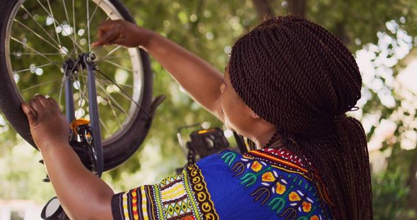 Zdravá černoška opravuje své vlastní kolo pomocí speciálního vybavení z nářadí na dvoře. Přibližovací záběr Dolly. Žena africký americký cyklista opravy přední kolo kolo venku. - Fotografie, Obrázek