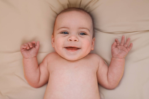 かわいい感情的な面白い笑顔の新生児の少年はベッドやベビーベッドにベージュシートに横たわっています. ベビーグッズの包装テンプレート。 健康な子供,病院の概念と幸せな母性. 幼い赤ちゃん。 ナースリー. - 写真・画像