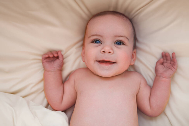 Roztomilý emocionální legrační úsměv novorozence ležící na béžových prostěradlech v posteli nebo postýlce. Šablona balení dětského zboží. Zdravé dítě, pojem nemocnice a šťastné mateřství. Dítě. Školka. - Fotografie, Obrázek