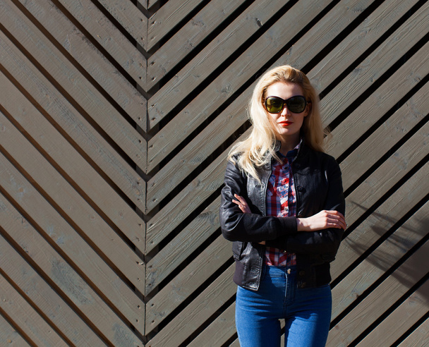 Beautiful blonde girl in huge sunglasses and a black jacket posing nex to wooden wall on a sunny day - Foto, Imagem