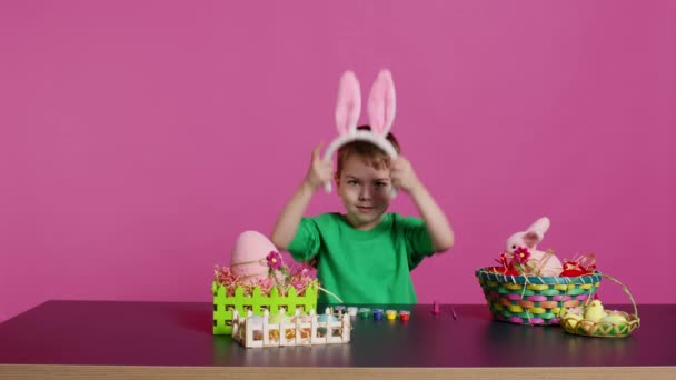 Sladké malé dítě, které si dává chlupaté králičí uši na hlavu a usmívá se před kamerou, šťastný nevinný chlapec sedící u stolu s uměleckými a řemeslnými potřebami na zdobení velikonočních vajíček. Jarní prázdniny. Fotoaparát B. - Záběry, video