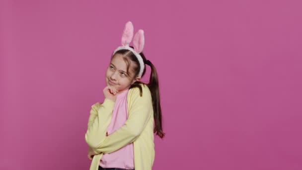 Glimlachend klein meisje dat droomt over geschenken en eten voor de paaszondag, enthousiast over de voorjaarsvakantie. Jonge jongen met konijnenoren poseren tegen een roze achtergrond. Camera A. - Video